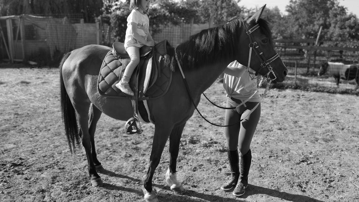 A grayscale image of a young girl riding a horse with a woman assisting on a farm.